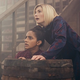 Mandip Gill and Jodie Whittaker as Yaz and the 13th Doctor, hiding behind a barrel as they stare in fear at something off-camera.